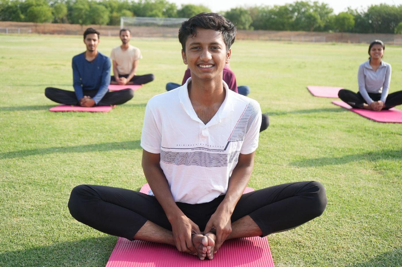 Navpratap Singh smiling in a seated yoga pose on a pink mat, with students practising behind him